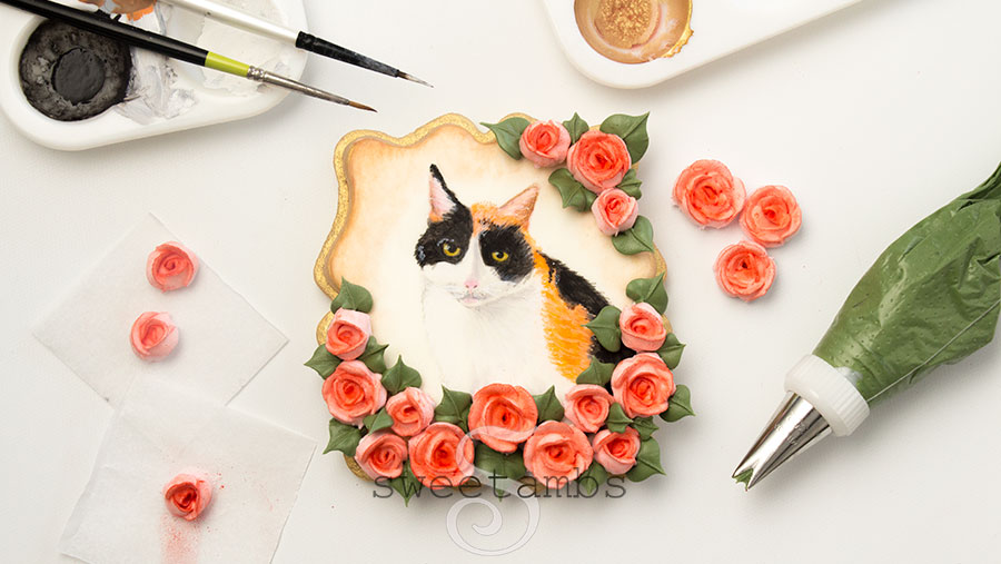 Calico cat cookie. This is a plaque shaped cookie with a painted calico cat surrounded by royal icing roses. The edge of the cookie is painted gold. There are royal icing roses scattered around the cookie. There is a piping bag filled with green icing next to the cookie. Above the cookie are two paint palettes with black, white, and gold edible paint as well as two small paint brushes sitting on top of the palette. 