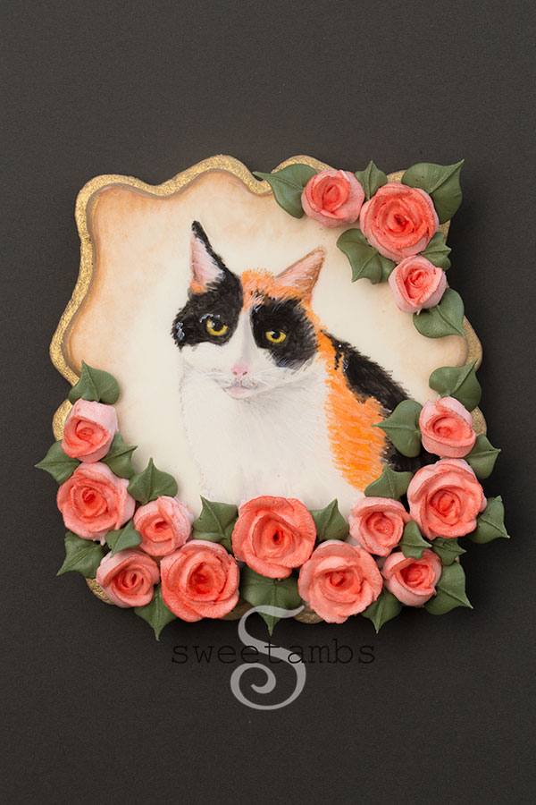 Calico cat cookie. This is a plaque shaped cookie with a painted calico cat surrounded by royal icing roses. The edge of the cookie is painted gold. 