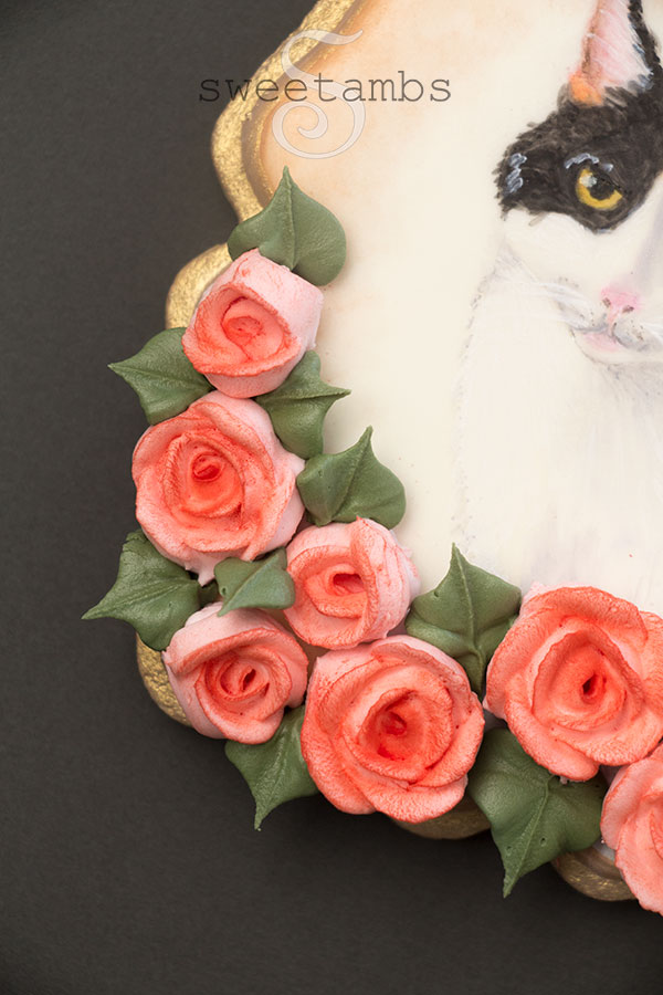 A close up of a calico cat cookie. This is a plaque shaped cookie with a painted calico cat surrounded by royal icing roses. The edge of the cookie is painted gold.