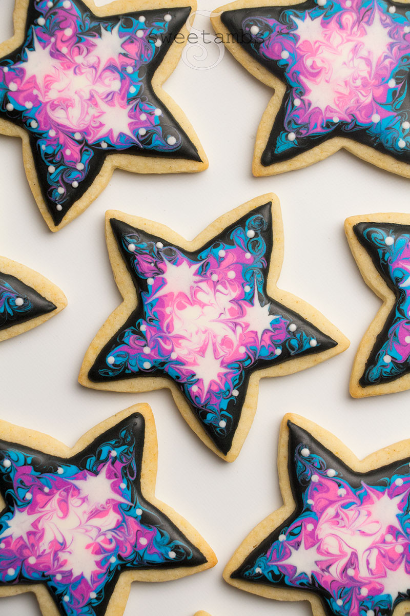 star shaped galaxy royal icing cookies decorated with black, blue, purple, pink, and white royal icing. The icing colors are swirled together. There are white royal icing stars in the base layer of icing. The cookies are on a white background.