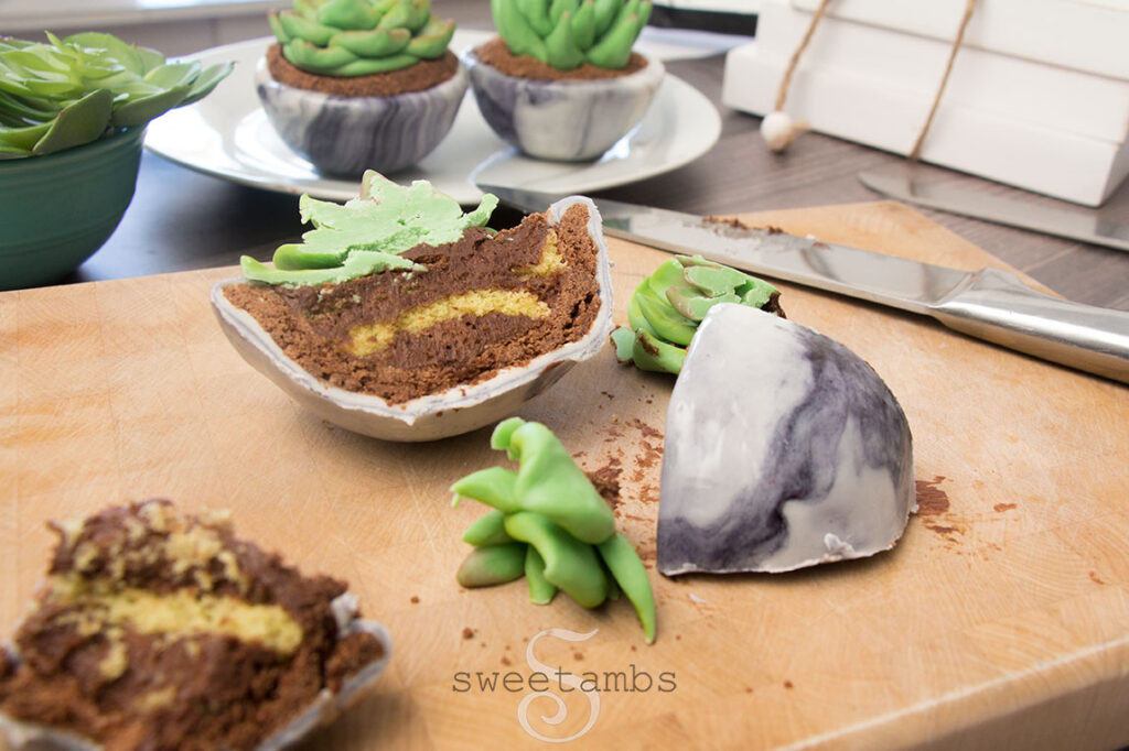 a cross-section of a succulent dessert. The succulent dessert is cut in half with pieces scattered on the cutting board. Inside the dessert are layers of chocolate frosting and yellow cake. There are more succulent desserts on a plate in the background and there is a knife on the cutting board. 
