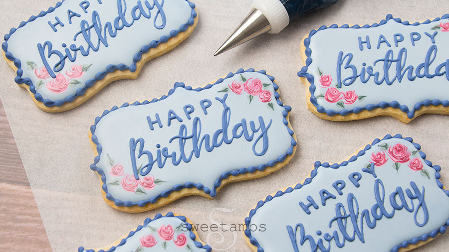 A set of plaque shaped cookies decorated with light blue royal icing and pink roses in the corners. Happy birthday is written on the cookies in royal icing. There is a piping bag filled with dark blue icing with a decorating tip. The cookies are on a piece of parchment paper on a wooden background.