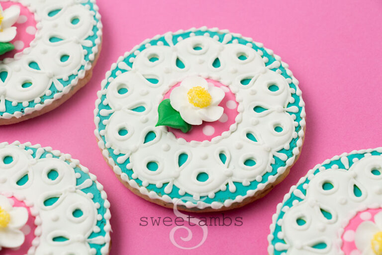 Round cookies decorated with teal royal icing and a white eyelet lace design. There is a pink and light pink pattern in circle in the middle of the lace. There is a royal icing strawberry blossom on top of the cookie. The cookies are on a bright pink background.