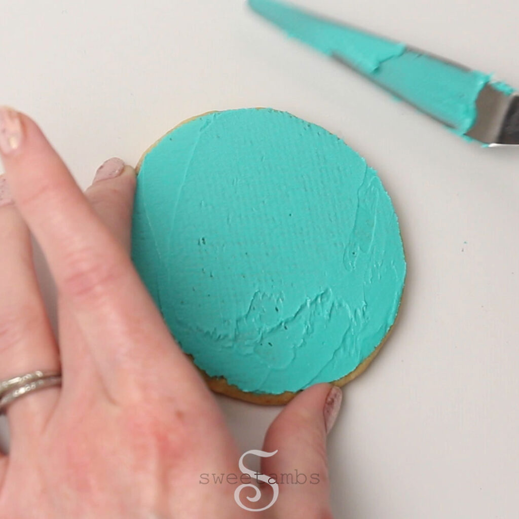A hand is hold a round cookie and a tapered spatula has just smoothed a layer of teal royal icing on the cookie.
