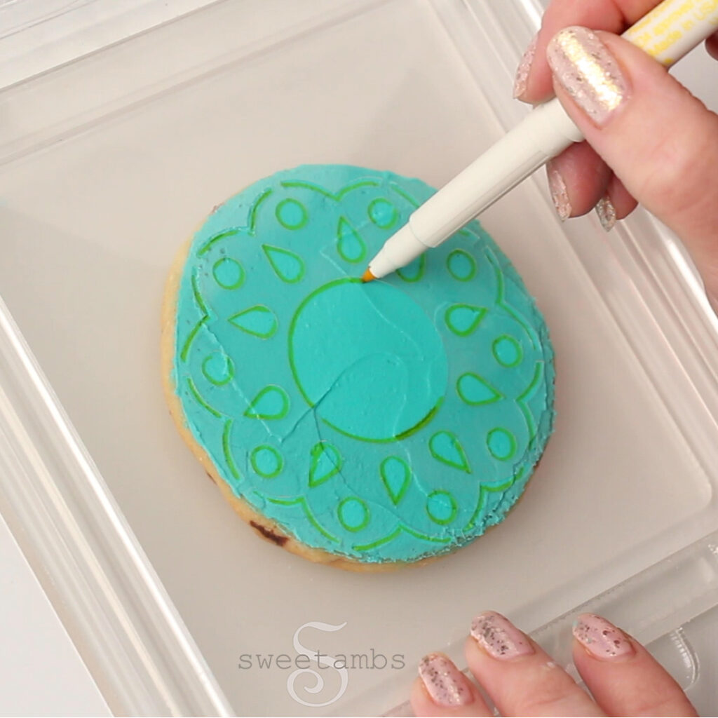 A hand holding an edible ink marker tracing an eyelet lace stencil on teal royal icing. the stencil is being held by a stencil holder.