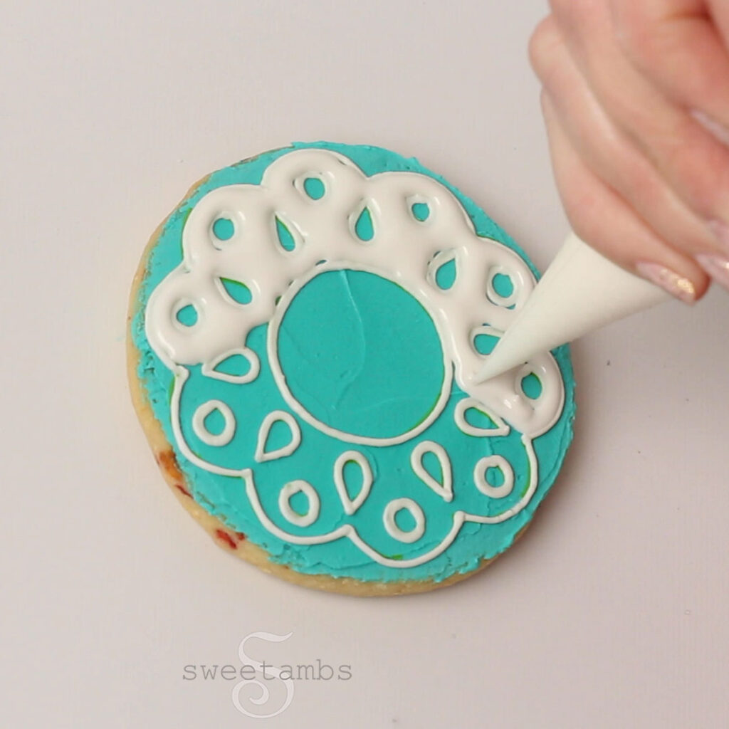 A hand holding a piping bag and filling in an eyelet lace design with white royal icing. The design is halfway filled in. The base layer of icing is teal green.