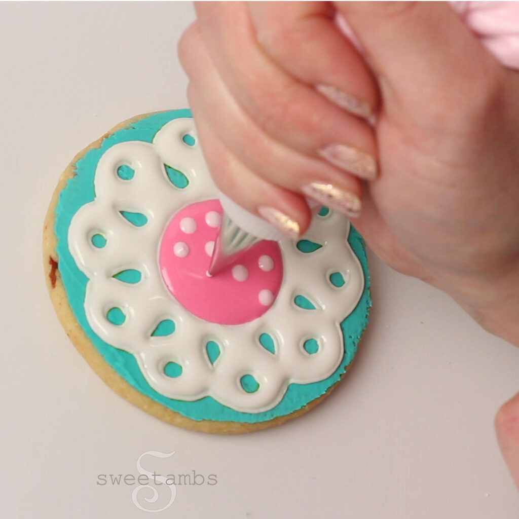 A hand piping light pink dots into a bright pink circle of icing in the center of the teal and white eyelet lace cookie.