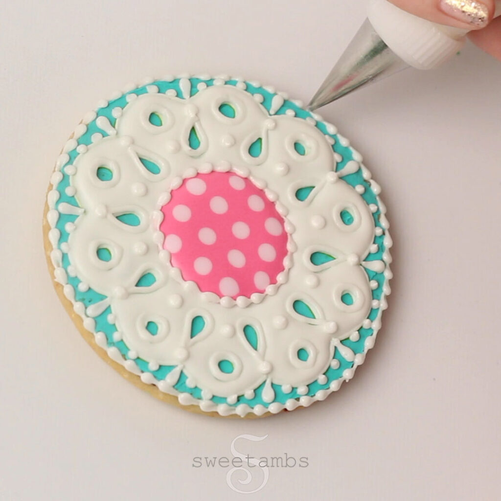A decorating tip adding details to the teal, white, and pink eyelet lace cookie