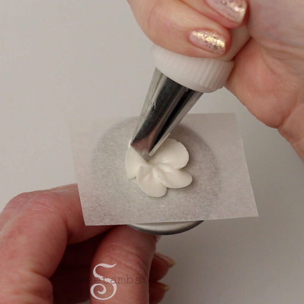 A hand holding a piping bag fitted with a petal tip and piping a strawberry blossom on a piece of parchment paper on a flower nail. The flower nail is being held by the other hand.