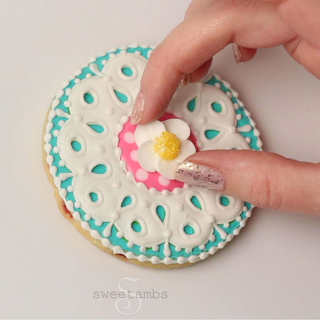 A hand with glitter nail polish placing the strawberry blossom in the center of the teal, white, and pink eyelet lace cookie.