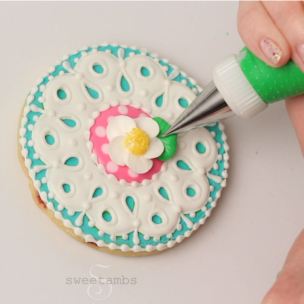 A hand holding a piping bag fitted with a leaf tip 352 piping a leaf on the strawberry blossom on the teal, white, and pink eyelet lace cookie.