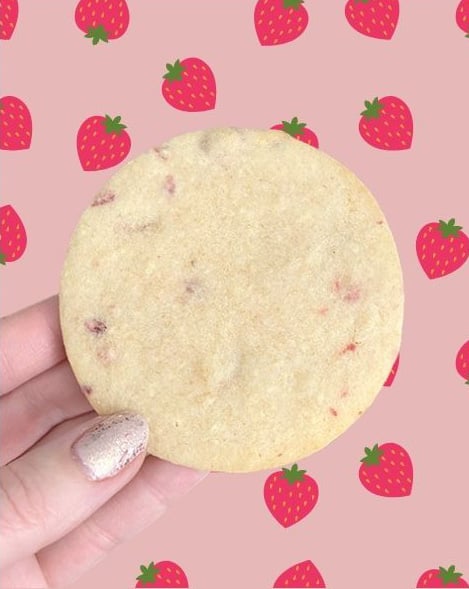 A round strawberry cookie being held by a hand with glitter nail polish. The background is pink with a red strawberry pattern.