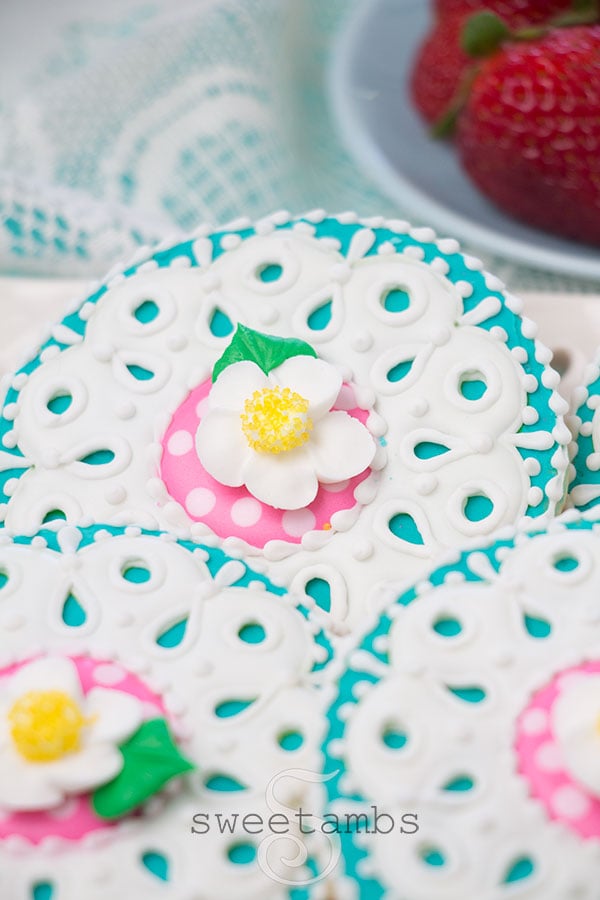 strawberry blossom cookies with royal icing eyelet lace decoration on a platter with an eyelet lace design. The platter is on a teal background to match the teal icing on the cookies. There is a lace fabric under the platter above the cookies. There is a light blue dish holding fresh strawberries in the background.