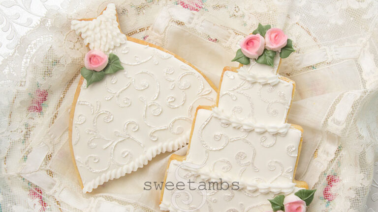 Wedding cookies decorated with royal icing filigree designs and royal icing roses. One cookie is shaped like a wedding gown and the other is shaped like a tiered wedding cake. The cookies are on piece of lace fabric on a floral platter.