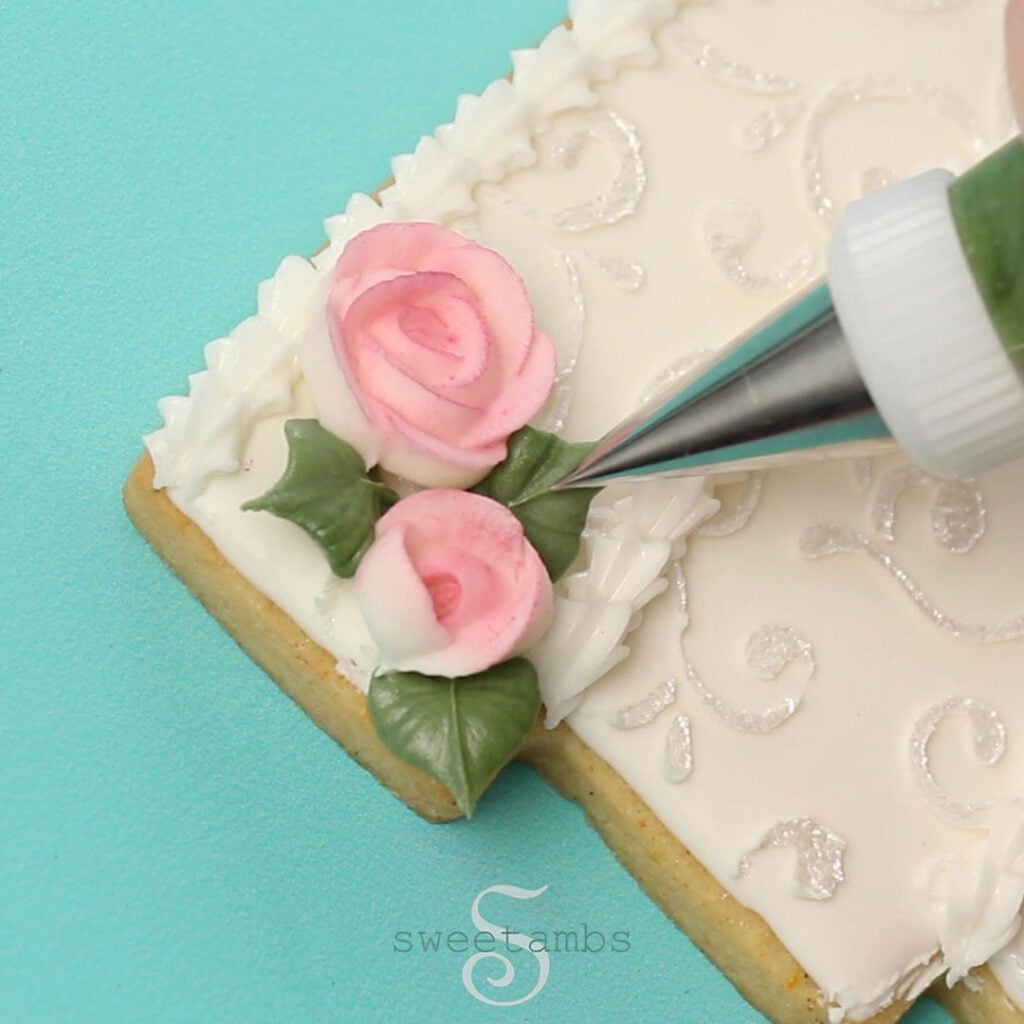 A decorating tip 352 is piping green royal icing leaves next to the royal icing roses on the tiered wedding cake cookie. The cookie is decorated with a shimmery filigree design and a shell border. The cookie is on a teal background.