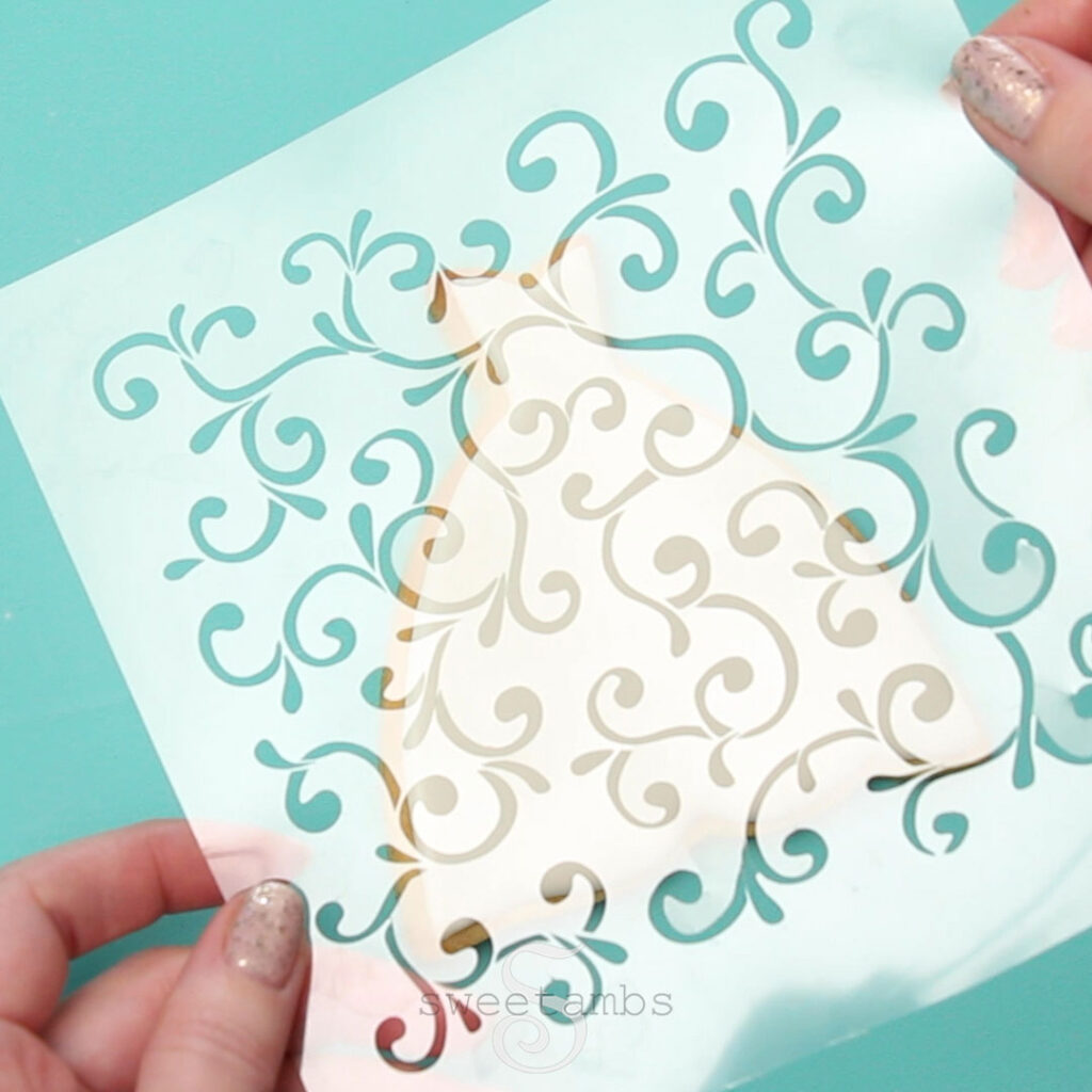 Hands holding a clear plastic filigree stencil. There is a wedding dress cookie on a teal background behind the stencil. 