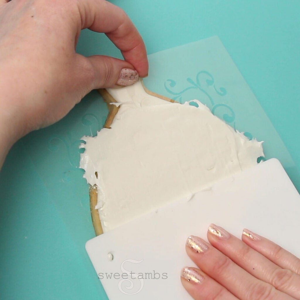 One hand is holding the filigree stencil on the wedding dress cookie while the other hand spreads royal icing over the stencil. The cookie is on a teal background.