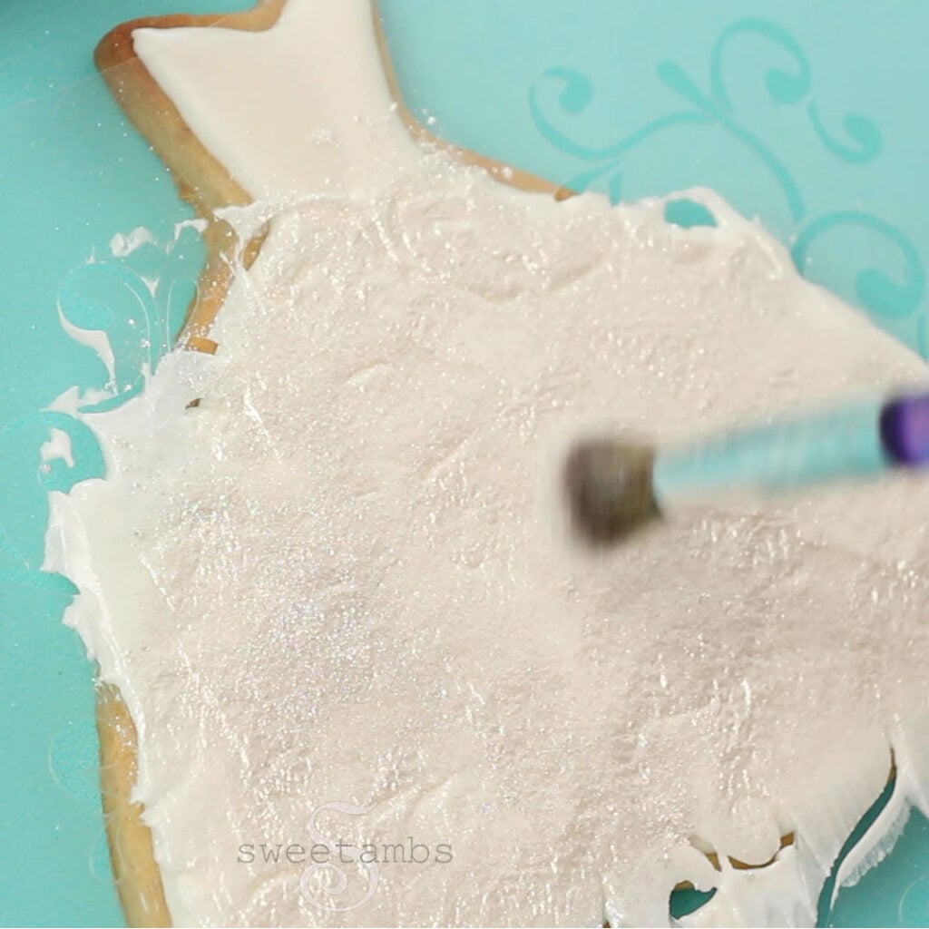 A brush is applying white pearl dust to the royal icing after it has been applied to the filigree stencil. The cookie is on a teal background.