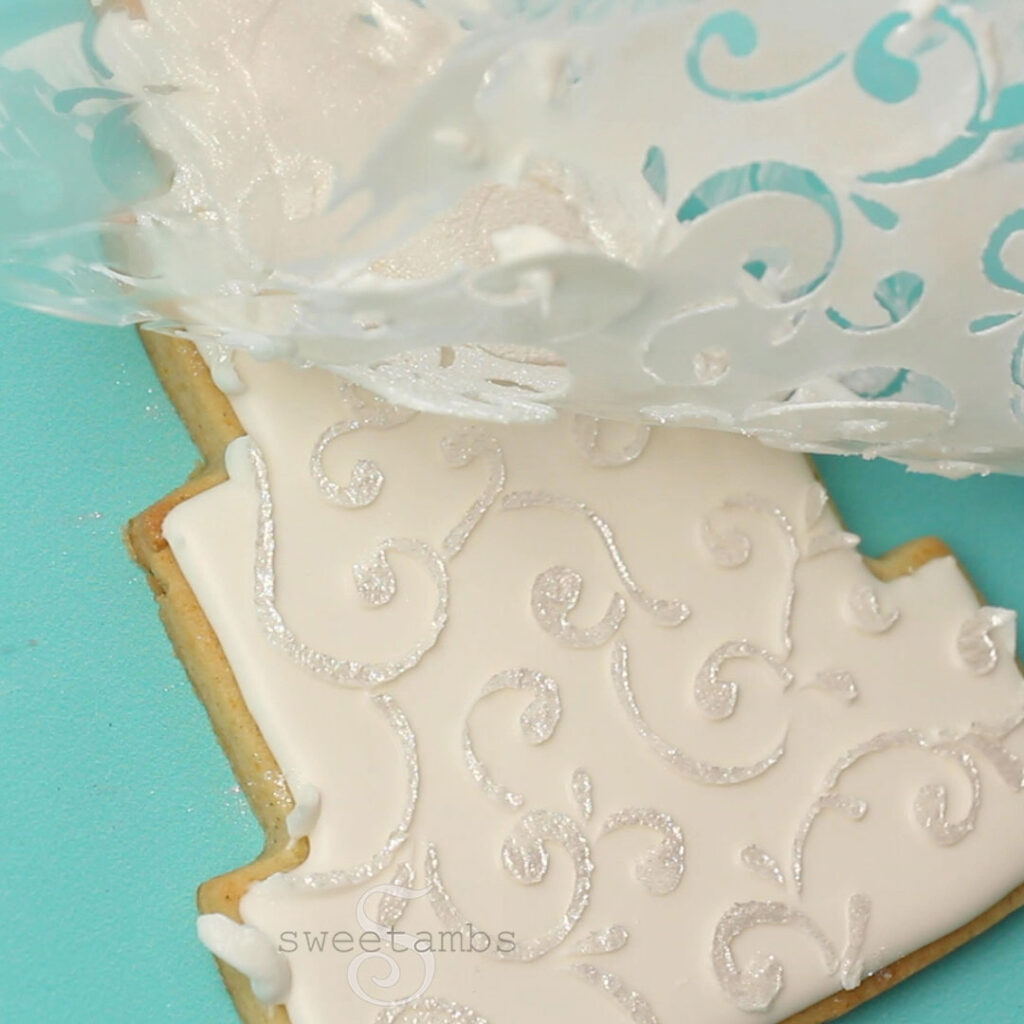 The stencil is being removed from the tiered wedding cake cookie revealing a shimmering filigree design. The cookie is on a teal background. 
