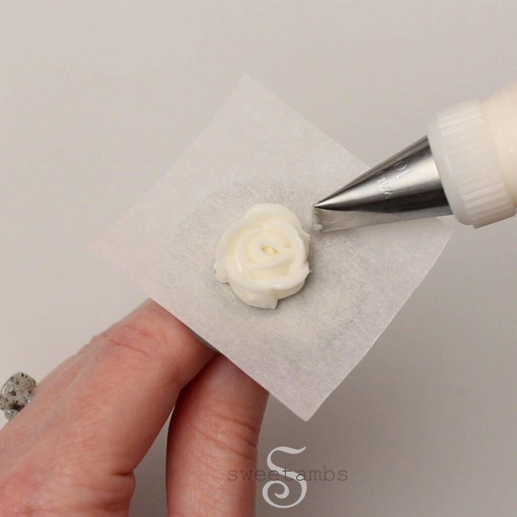 A hand is holding a flower nail with a parchment square. There is a decorating tip 101 piping a white royal icing rose on the parchment square. 
