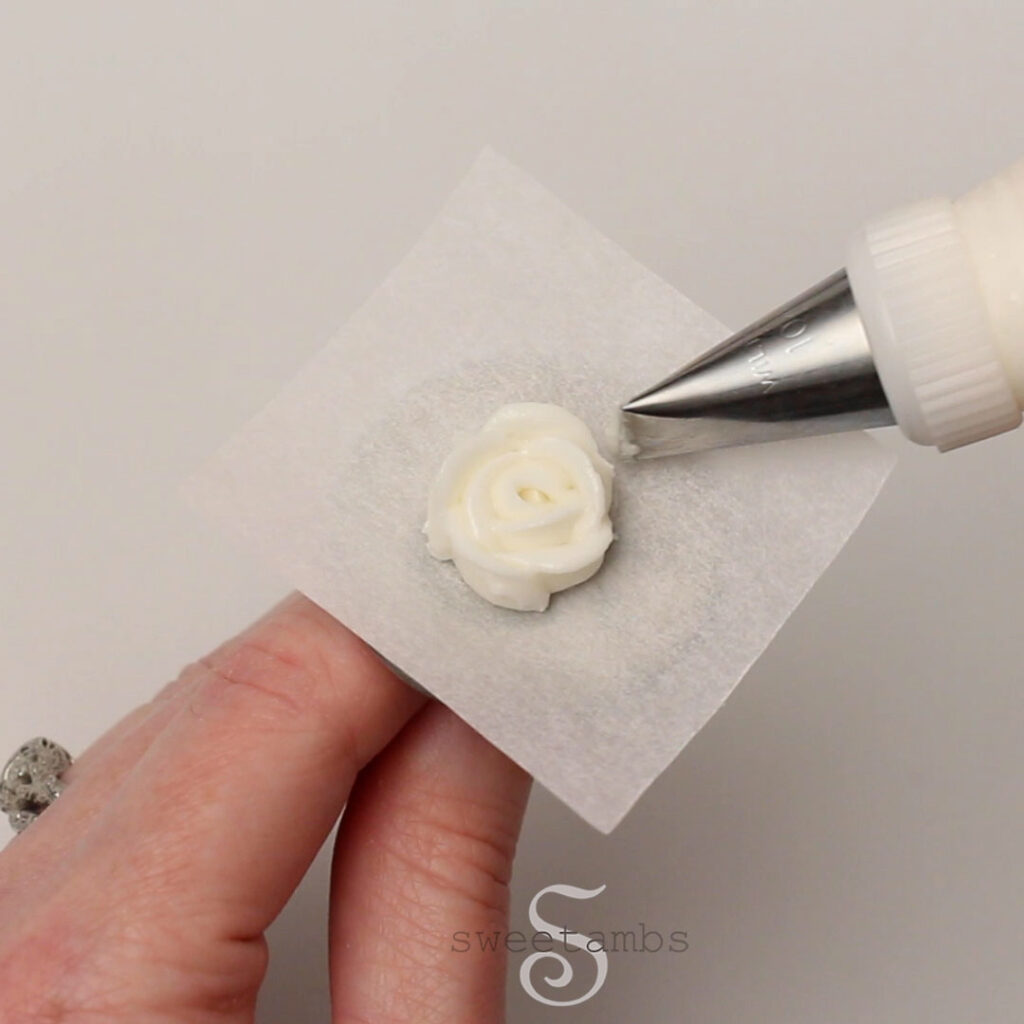A hand is holding a flower nail with a parchment square. There is a decorating tip 101 piping a white royal icing rose on the parchment square.
