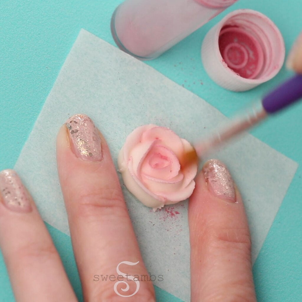A brush is applying pink color dust to the royal icing rose. There are 3 fingers with glitter nail polish holding the rose in place. There is an open container of pink dust above the rose. There is a teal background.