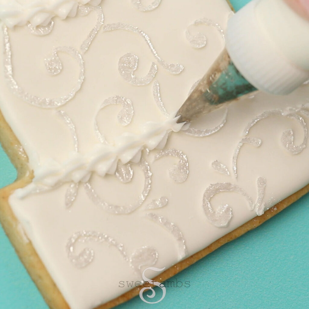 A decorating tip 16 is piping a shell border on the tiered wedding cake cookie. The cookie is decorated with a shimmery filigree design. The cookie is on a teal background. 