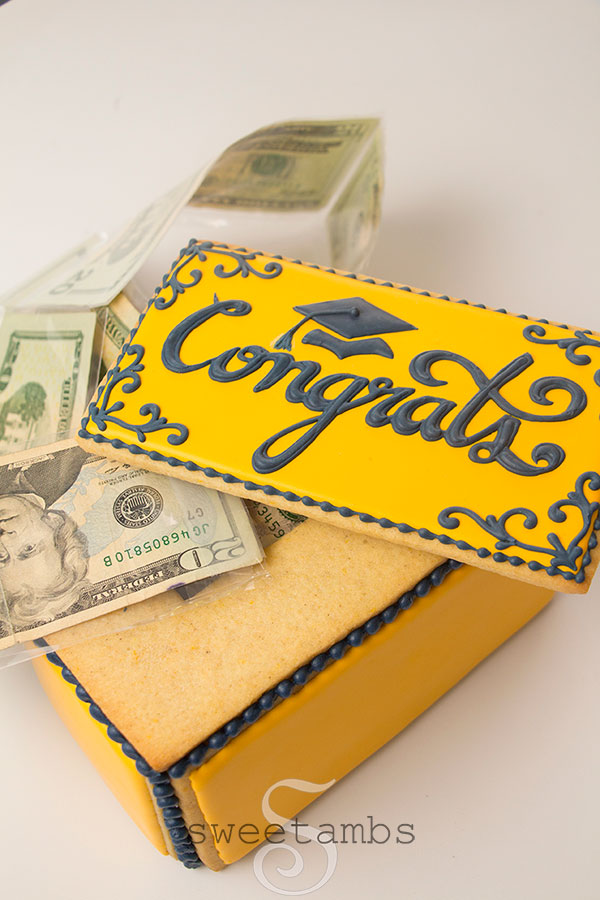 a cookie box with money coming out of it. The top of the box is askew. It is decorated in royal icing with the word "congrats" and a graduation cap.