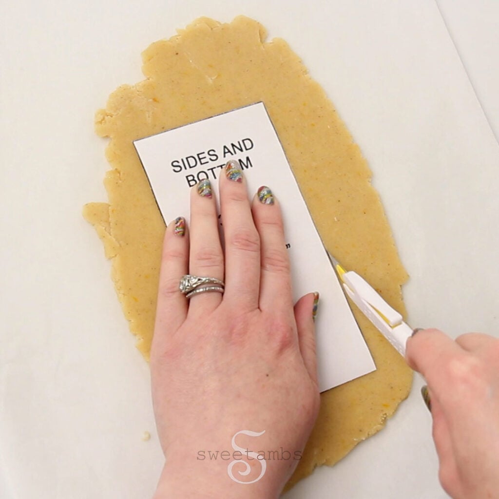 A hand holding a rectangular paper template on a sheet of cookie dough while the other hand cuts around the template with a craft knife.