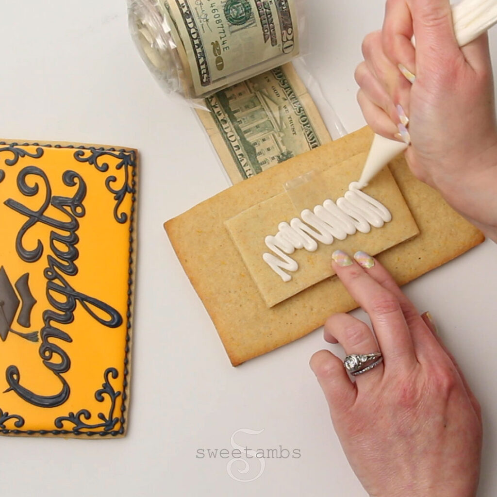 A hand is holding a small cookie piece on top of a larger cookie piece. The other hand is piping white icing on the cookie. The  roll of money is underneath the lid and the decorated part of the lid is on the left side. The decorated cookie is golden yellow with the word "congrats" written in cursive.