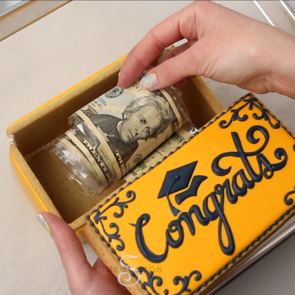 A hand is placing the roll of money inside the cookie box while the other hand holds the lid. The lid of the box is decorated with golden yellow royal icing with the word "congrats" written in cursive and there is a graduation cap above the word. The corners are decorated with filigree designs. The fingernails are painted with a pastel plaid design. 