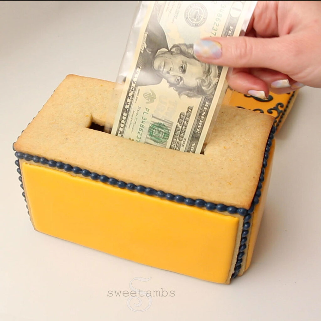a hand pulling a $20 bill out of the box. The nail on the thumb is painted with a pastel plaid design.