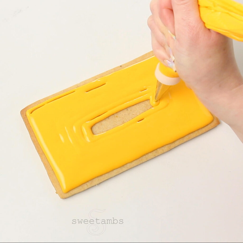 A hand holding a decorating bag and icing a rectangle cookie with golden yellow royal icing. The decorating bag has a metal decorating tip and a coupler.