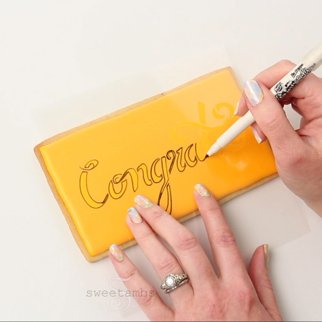 One hand is holding a "congrats" stencil on the golden yellow iced cookie while the other hand traces the stencil with a black edible ink marker. The fingernails are painted with a pastel plaid design.