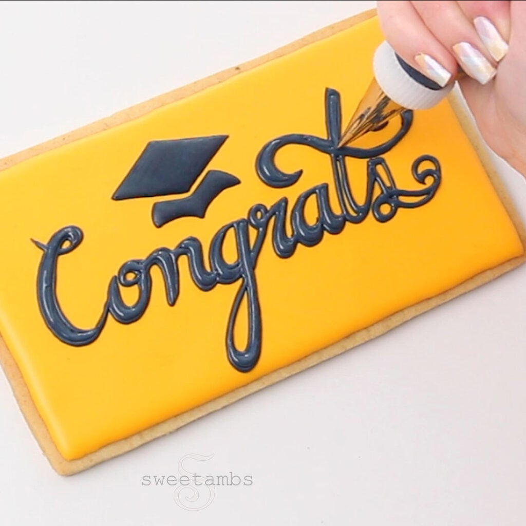 A hand is holding a piping bag fitted with a metal tip and a coupler and filling in the stenciled lettering with dark blue royal icing. The word "congrats" is written in cursive and there is a graduation cap above the word. The fingernails are painted with a pastel plaid design. 