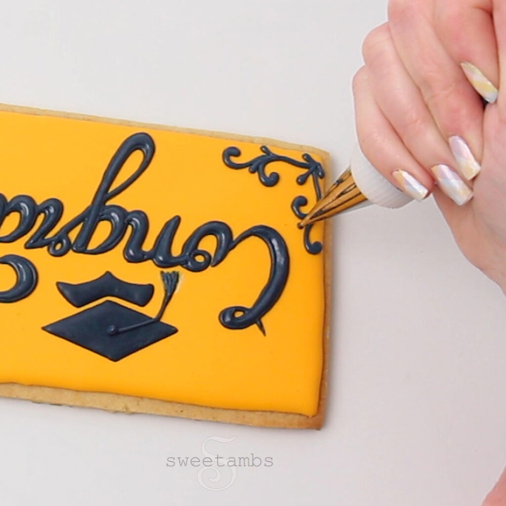 A hand is holding a piping bag fitted with a metal tip and a coupler and piping a filigree design on the corner with dark blue royal icing. The word "congrats" is written in cursive and there is a graduation cap below the word. The fingernails are painted with a pastel plaid design.