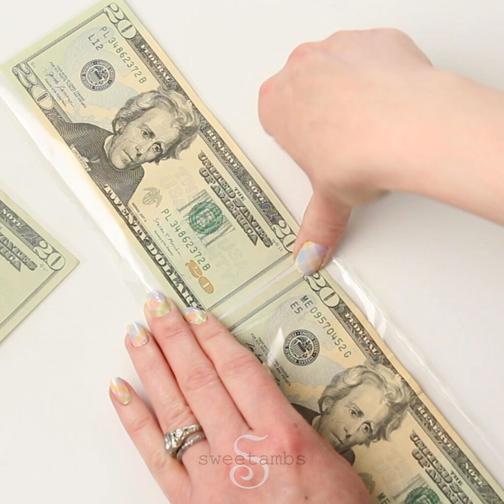 A hand is holding $20 bills together while the other hand applies tape between them. The bills are wrapped in cello bags. The fingernails are painted with a pastel plaid design. 