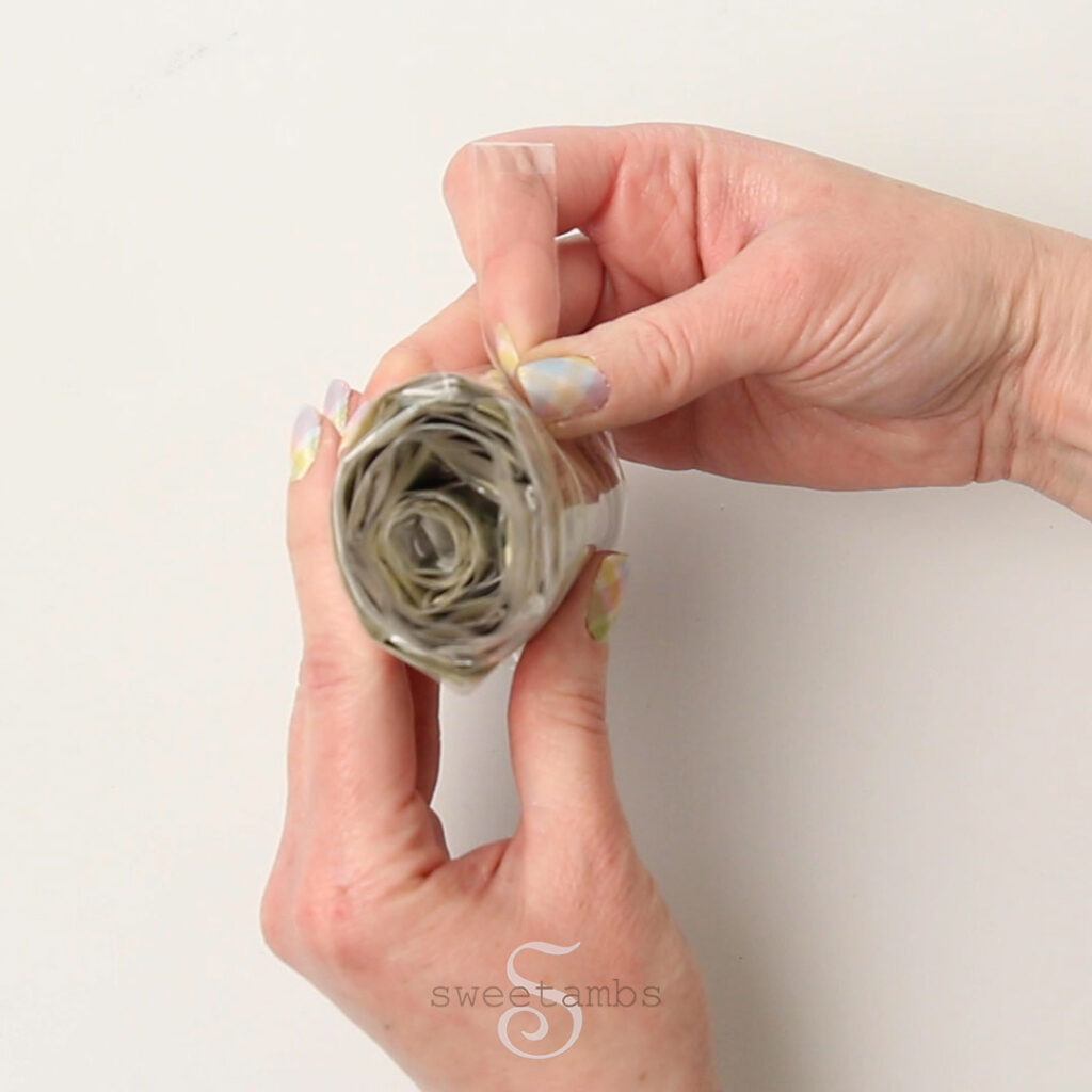 Two hands holding a roll of money wrapped in cello bags. The thumb and forefinger are holding a plastic tap at the end of the roll. The fingernails are painted with a pastel plaid design.