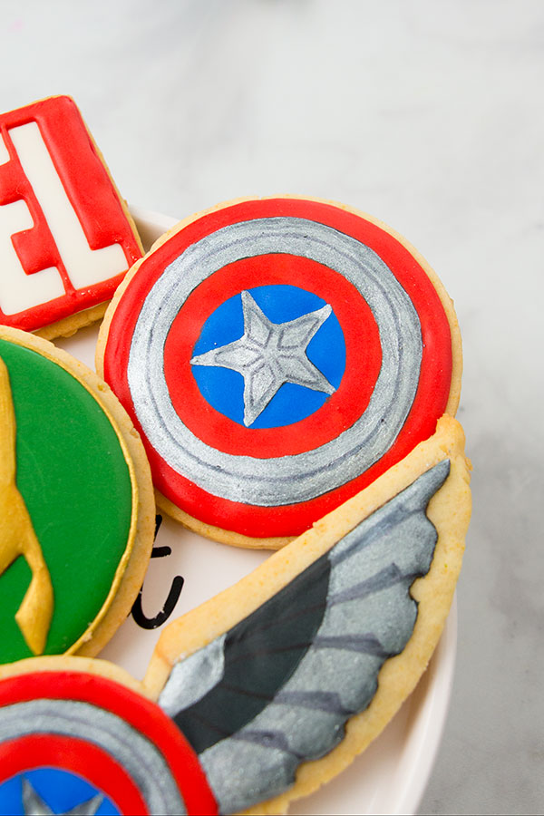 A close up of the Captain America shield cookie. The cookie is on a cake plate surrounded by other Marvel theme cookies.