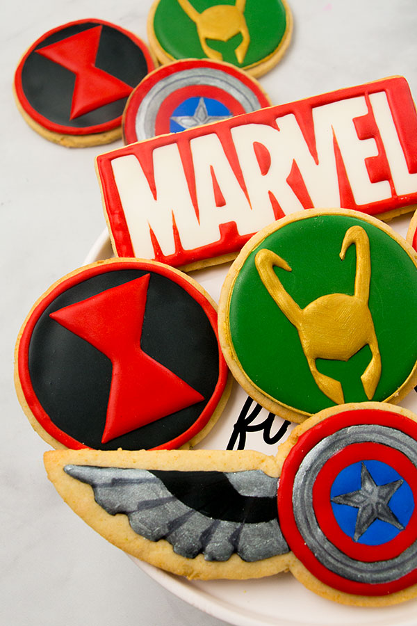 A set of marvel cookies on a cake plate. The cookies feature the Captain America Shield, Falcon's wings, the Black Widow emblem, and Loki's mask. There is a cookie in the group with the Marvel logo. There are 3 cookies in the background on a marble surface.