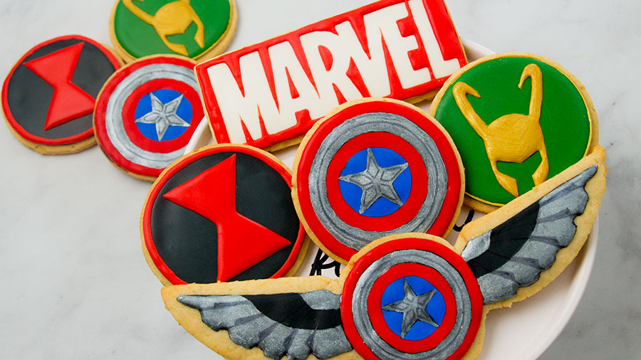 A set of marvel cookies on a cake plate. The cookies feature the Captain America Shield, Falcon's wings, the Black Widow emblem, and Loki's mask. There is a cookie in the group with the Marvel logo. There are 3 cookies in the background on a marble surface.