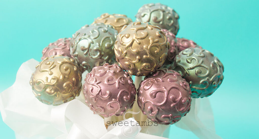 A bunch of shimmer cake pops in a cup with a puffy white ribbon surrounding the cup. The cake pops are decorated with a filigree design and dusted with gold, pink, and blue luster dust to make them shine.