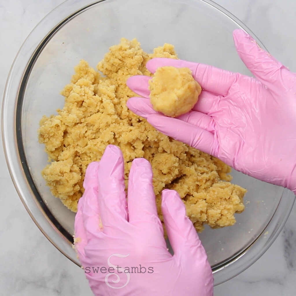  A pink gloved hand holding a ball of the cake pop mixture. The rest of the mixture is in a glass bowl. The other pink gloved hand is resting on the edge of the bowl