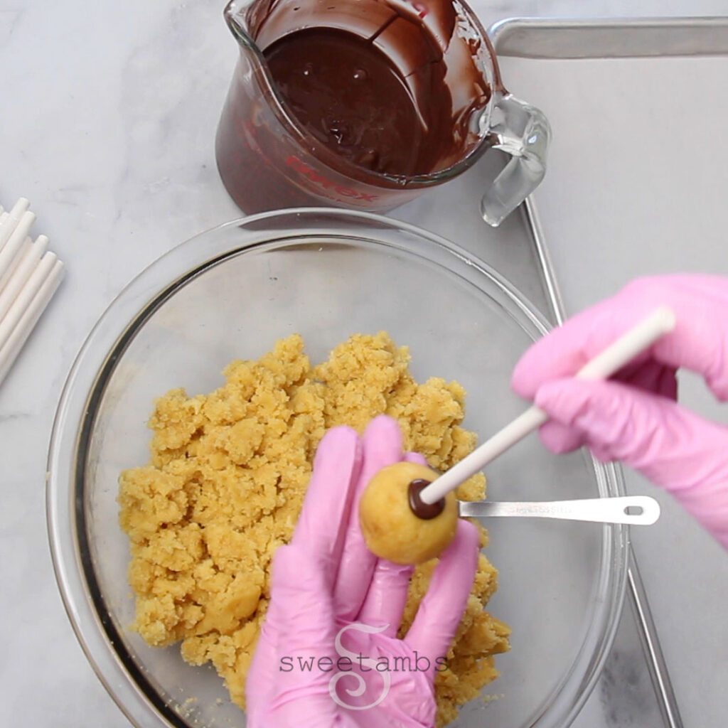 Pink gloved hands are inserting a lollipop stick that has been dipped in melted chocolate into a ball of the cake pop mixture. The rest of the cake pop mixture is in a glass bowl. There are lollipop sticks to the left of the bowl and a glass measuring cup full of melted chocolate above the bowl. There is a parchment lined baking sheet to the right of the bowl. 