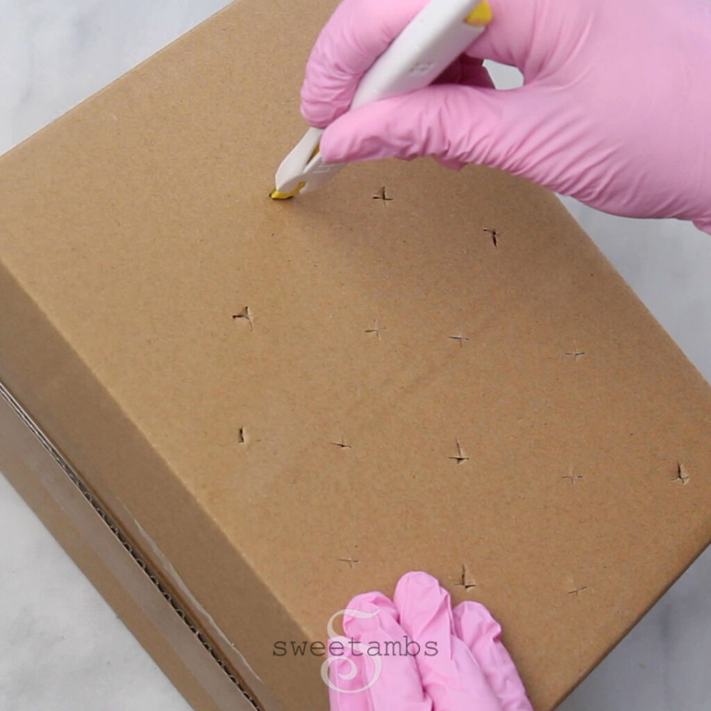 A pink gloved hand is using a craft knife to cut X shapes into a cardboard box to be used as a cake pop stand.