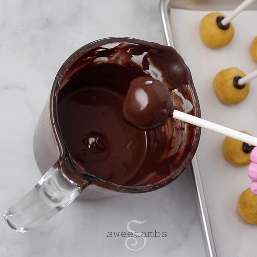 A chocolate coated cake pop being lifted out of a measuring cup full of melted chocolate. There is a parchment lined baking sheet to the right of the measuring cup and there are un-dipped cake pops lined up on the tray.