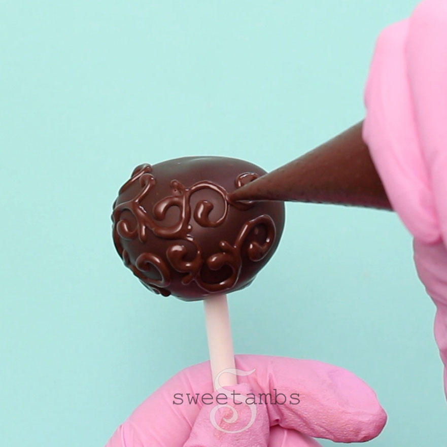 A pink gloved hand is piping a filigree design onto the cake pop. Another pink gloved hand is holding the lollipop stick.