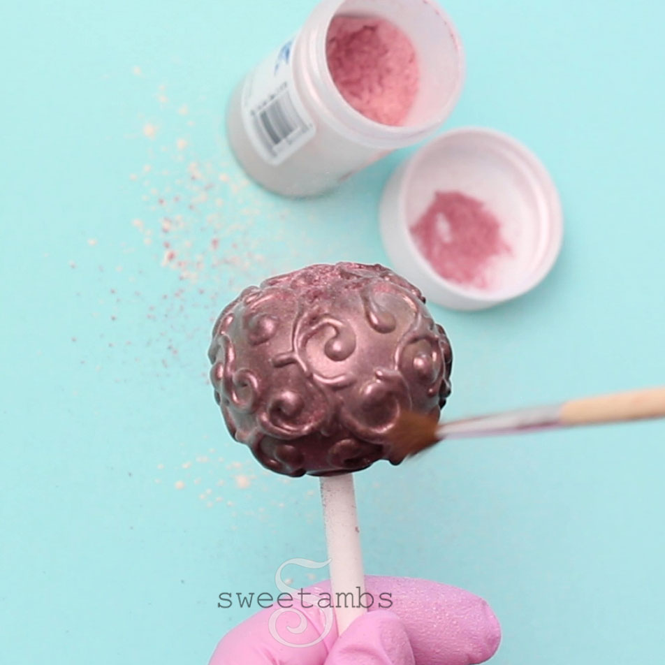 A brush is applying pink luster dust to the filigree decorated cake pop. There is an open container of pink luster dust in the background with some luster dust in the lid. There is a pink gloved hand holding the cake pop stick. 