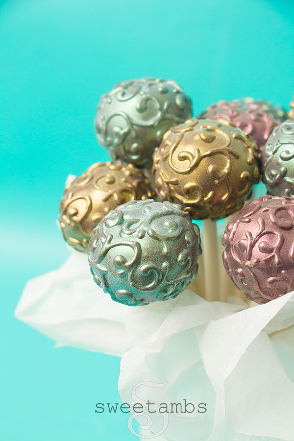 A bunch of shimmer cake pops in a cup with a puffy white ribbon surrounding the cup. The cake pops are decorated with a filigree design and dusted with gold, pink, and blue luster dust to make them shine.
