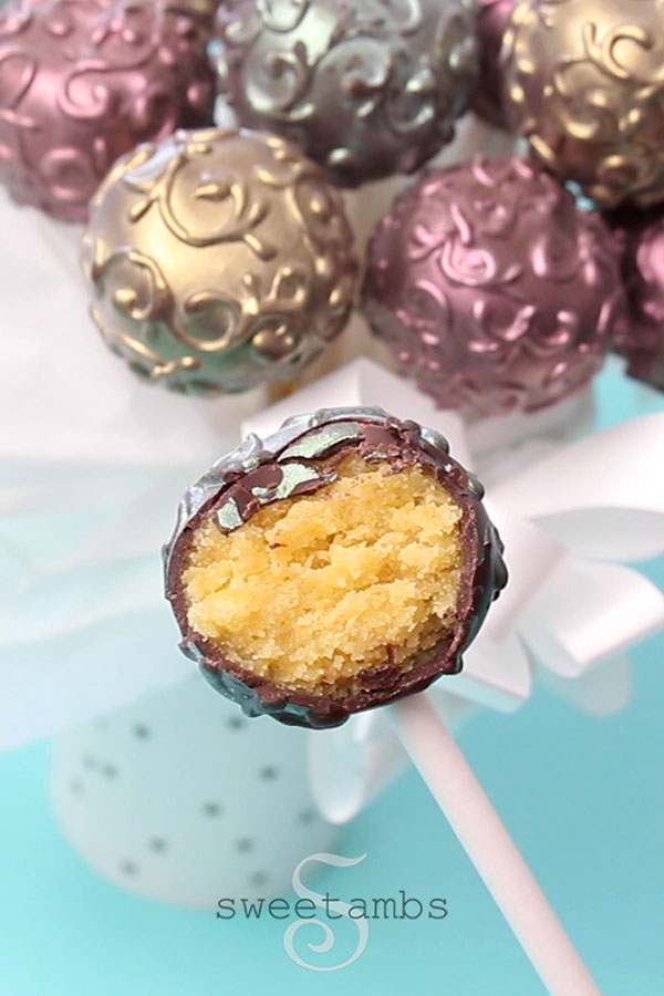 A cake pop with a bite taken out of it. In the background are a bunch of shimmer cake pops in a cup with a puffy white ribbon surrounding the cup. The cake pops are decorated with a filigree design and dusted with gold, pink, and blue luster dust to make them shine.